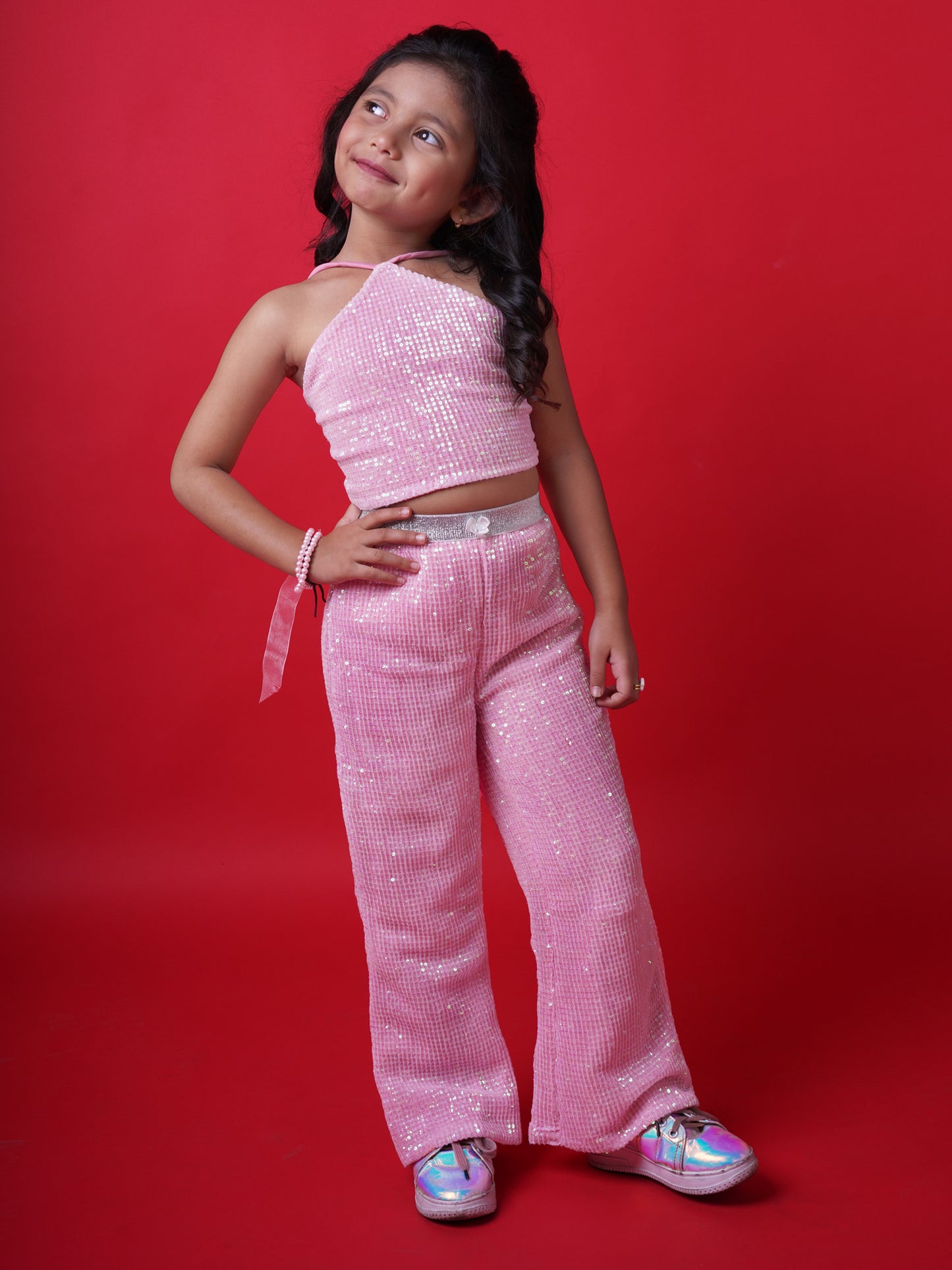 Young girl wearing a Little Carrot pink sequin halterneck top and matching flared pants, smiling with arms raised in a playful pose, standing against a red background.