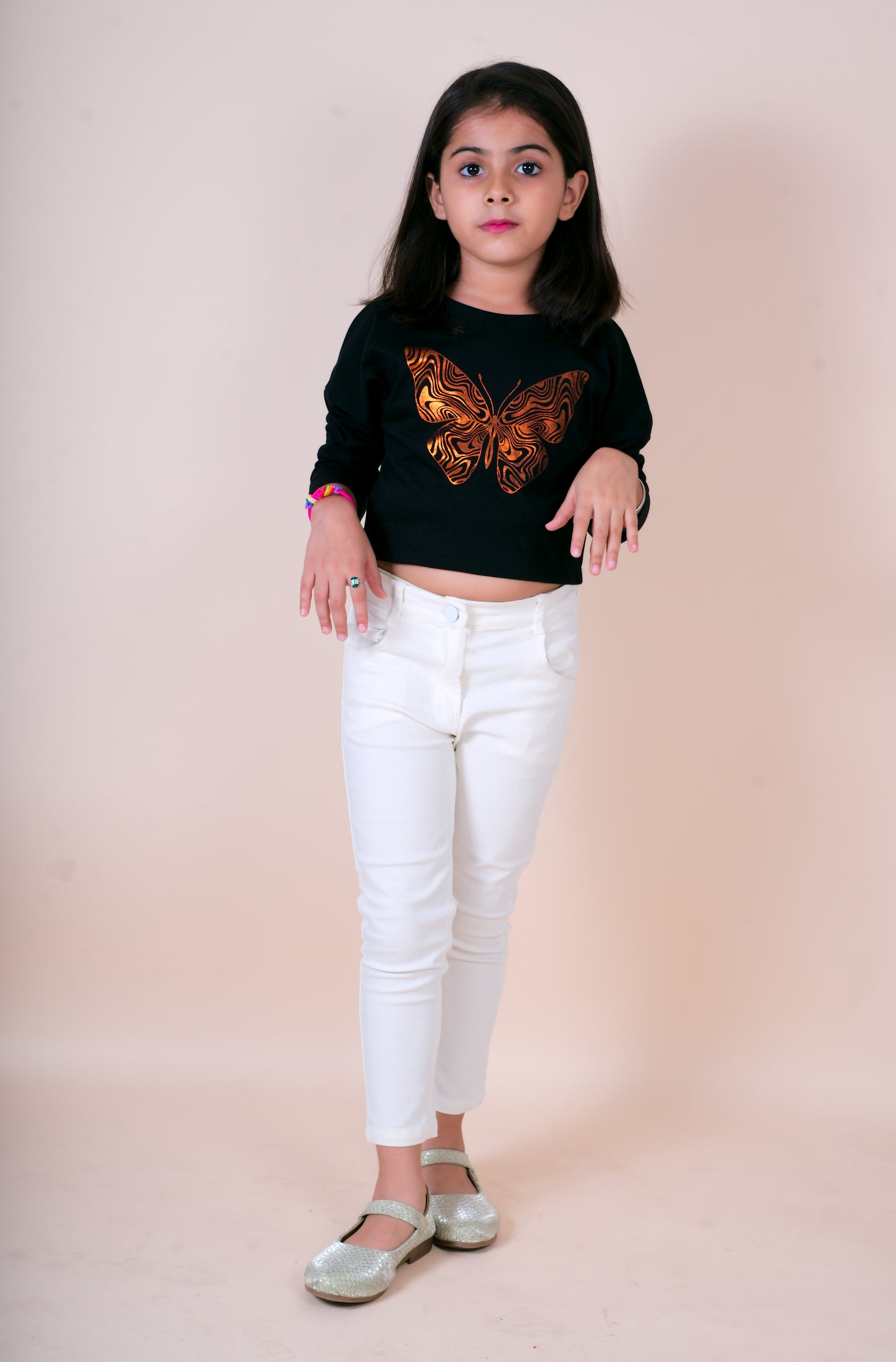 Young girl wearing a black sweater with a butterfly design and white pants on a beige background