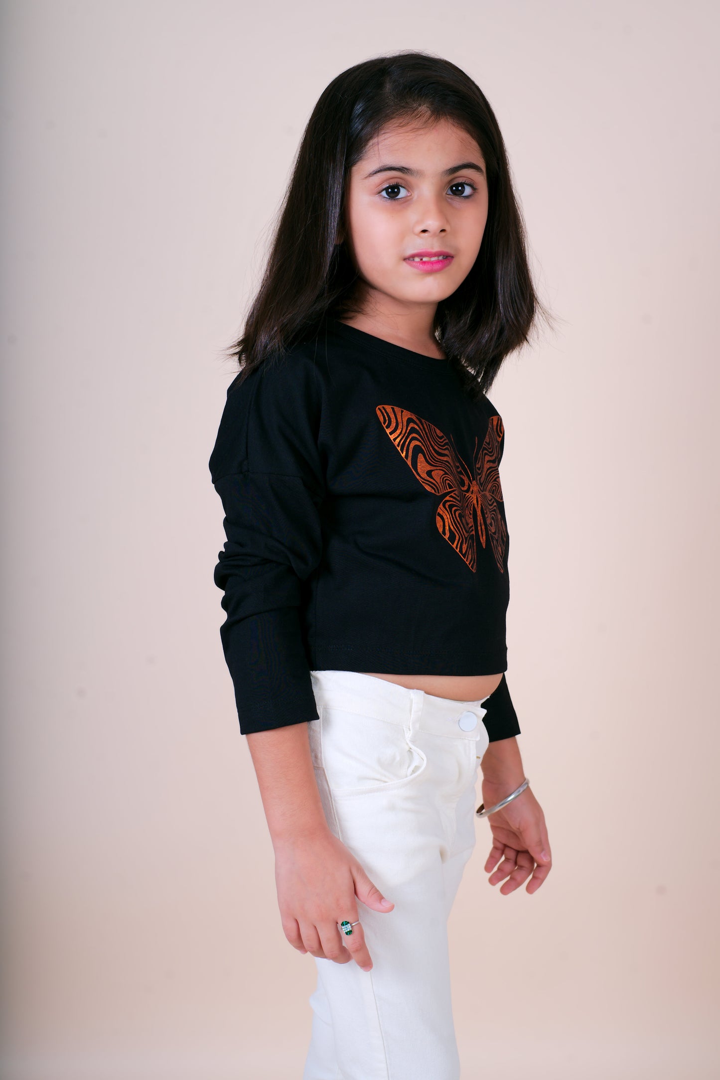 Young girl wearing a Little Carrotblack top with orange design and white pants against a plain background