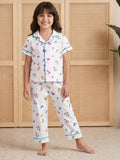 A young girl smiling while wearing Little Carrot two-piece white pajama NIght Suit set with a colorful butterfly print and light blue trim, standing in front of a beige woven cabinet.