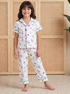 A young girl smiling while wearing Little Carrot two-piece white pajama NIght Suit set with a colorful butterfly print and light blue trim, standing in front of a beige woven cabinet.