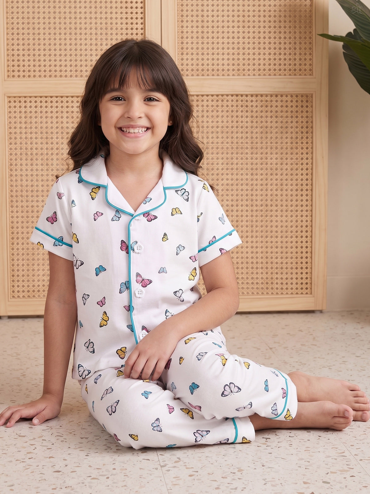 A young girl with dark brown hair sits smiling on a light speckled floor, wearing Little Carrot white short-sleeve pajama Night Suit set with colorful butterfly prints and light blue piping. She is posed in front of a beige cane-screen cabinet.