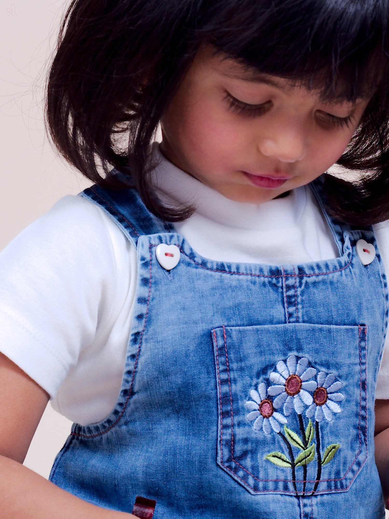 A stylish denim overall dress for infant and toddler girls, featuring floral embroidery on the front pocket, adjustable buttoned straps, and a ruffle hem for a cute and comfortable look.