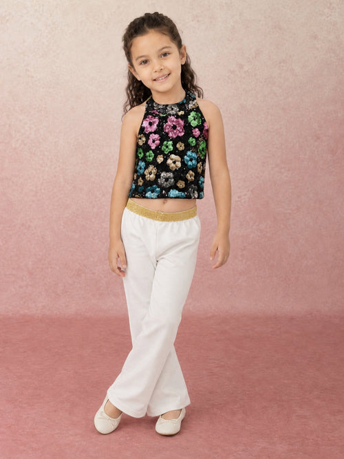 A young girl modeling a sleeveless black sequined crop top with colorful floral sequins from pink skies collection by Little Carrot, paired with white pants and white shoes, standing against a textured pink backdrop.