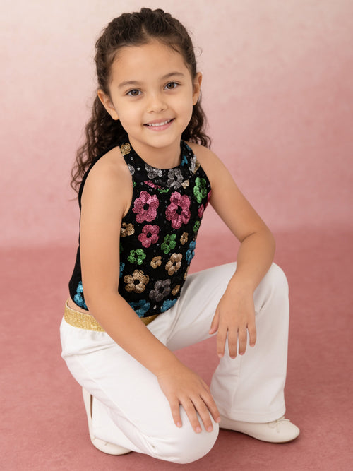 A young girl modeling a sleeveless black sequined crop top with colorful floral sequins from pink skies collection by Little Carrot, paired with white pants and white shoes, standing against a textured pink backdrop.
