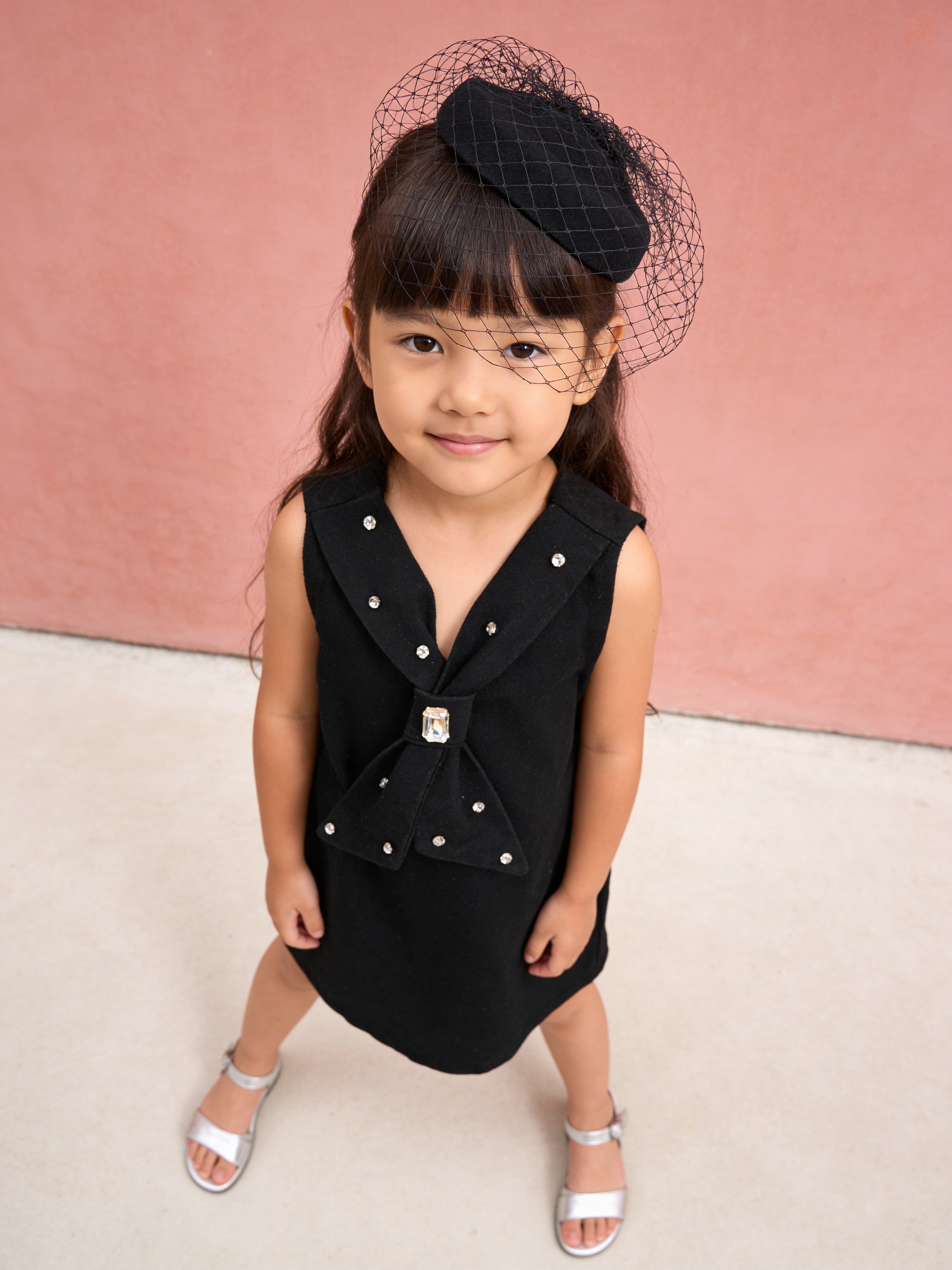 Young girl wearing a Little Carrot black Denim dress with a large bow with hand  Stone Embellsigment and matching headband against a pink background