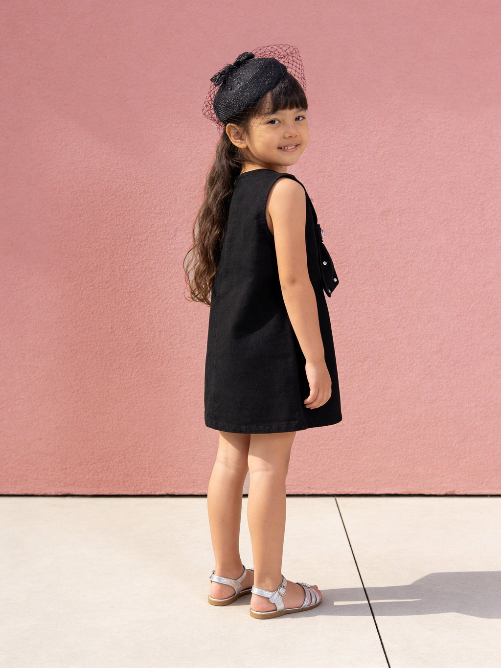 Young girl in a little carrot black denim dress standing against a pink wall