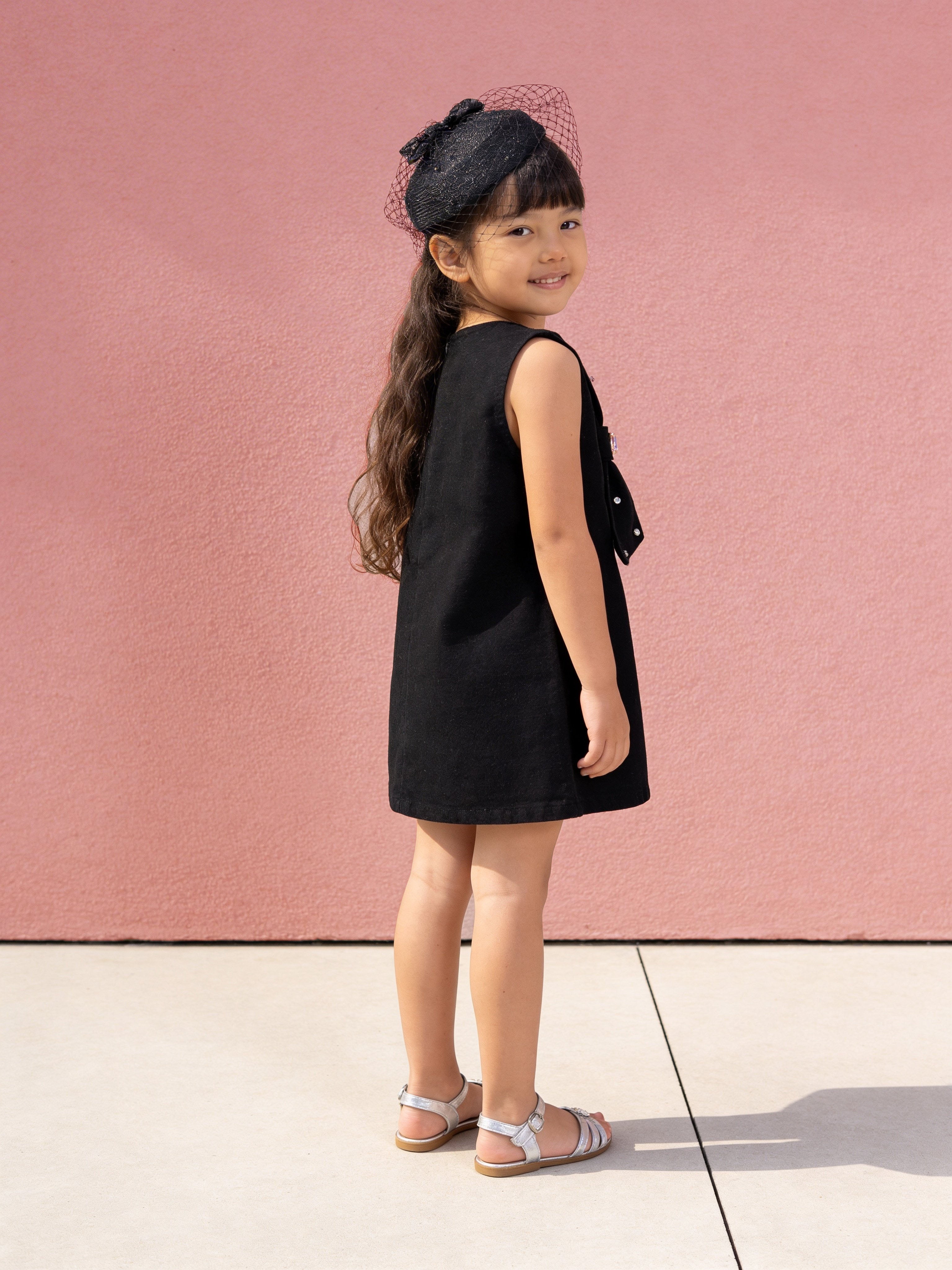 Young girl in a little carrot black denim dress standing against a pink wall