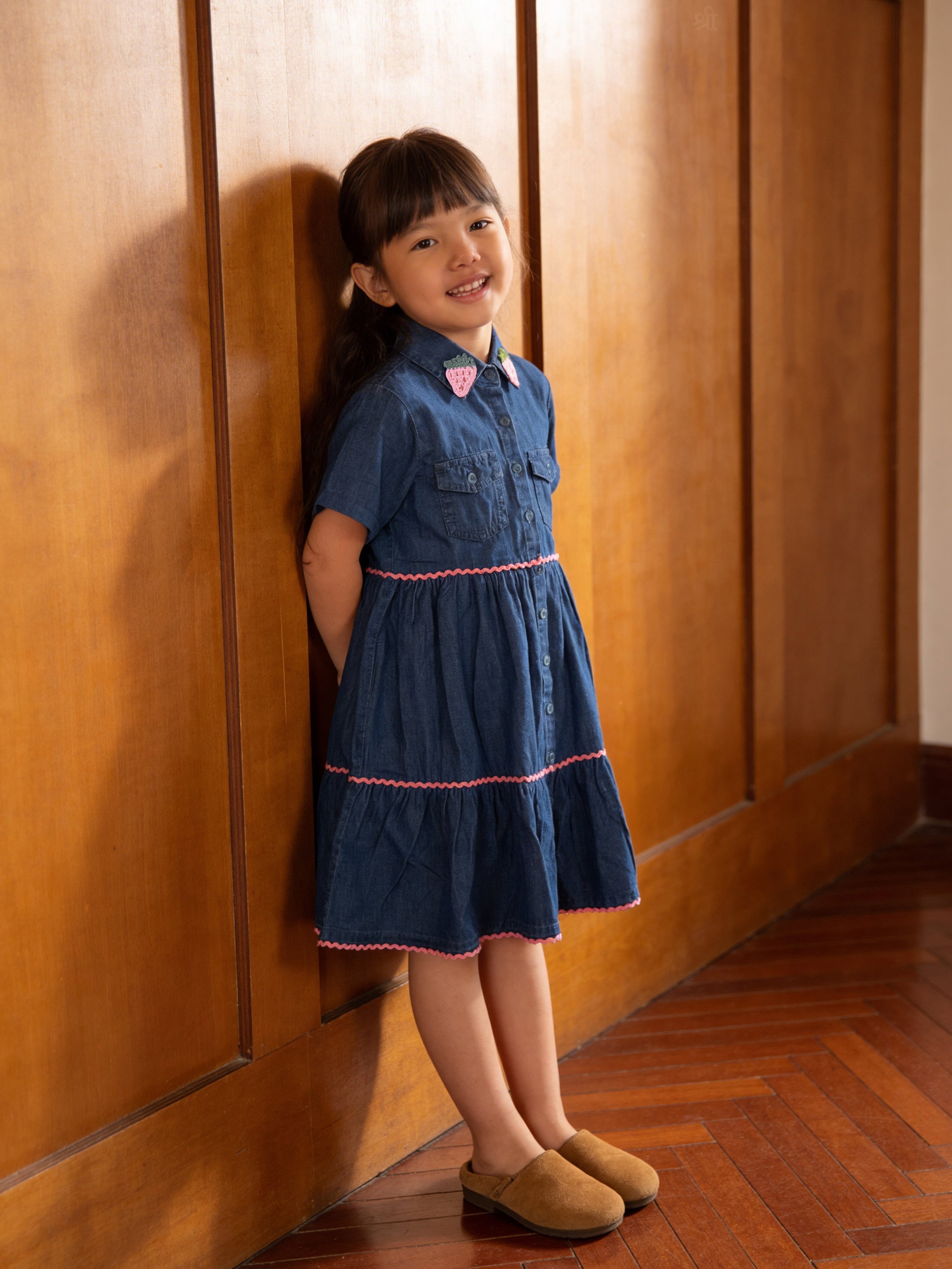 Young girl in a Little Carrot denim dress standing against a wooden wall.