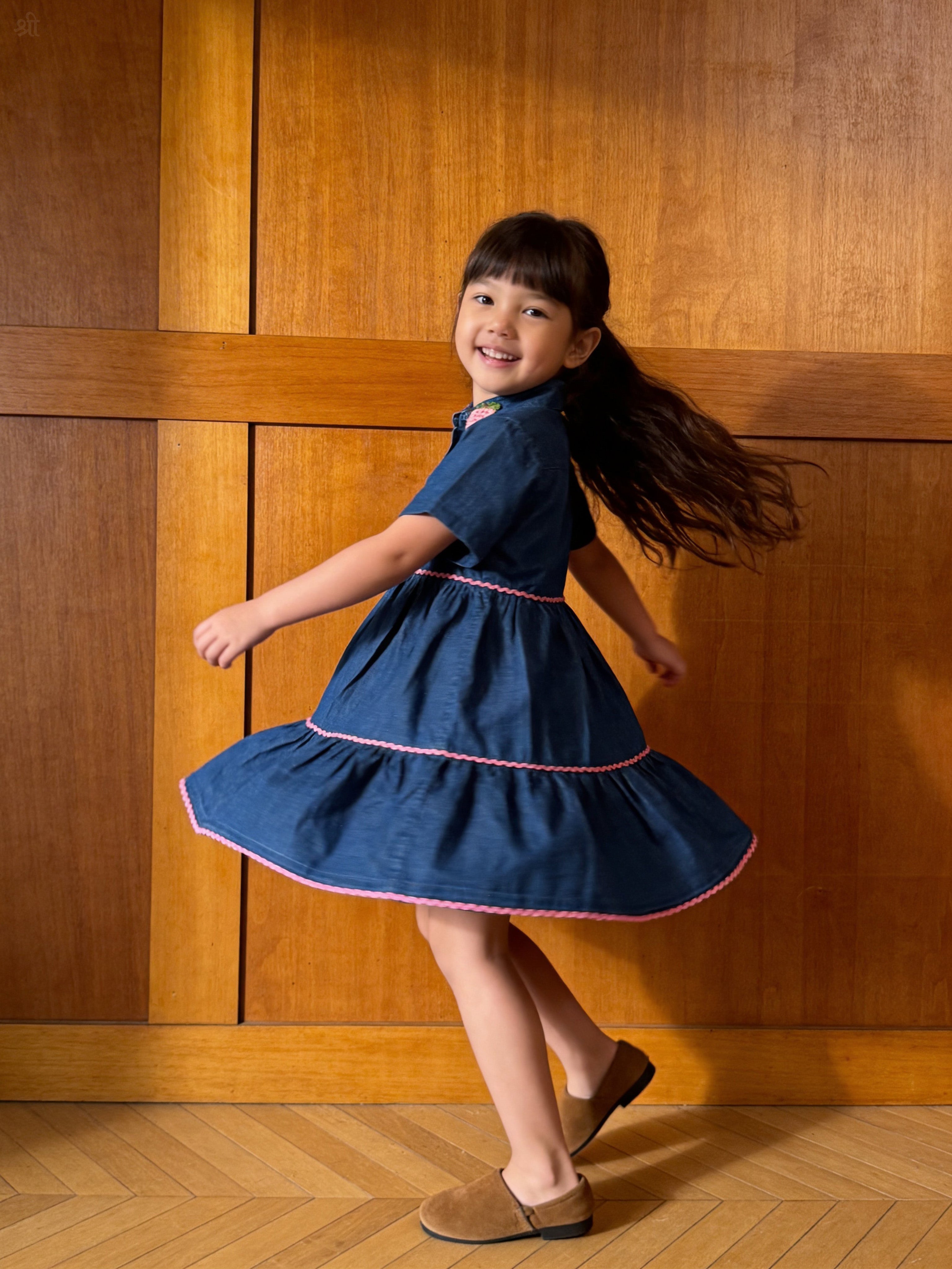 Young girl in a Little Carrot blue denim dress with pink hand crochet trim dancing in front of wooden paneling.
