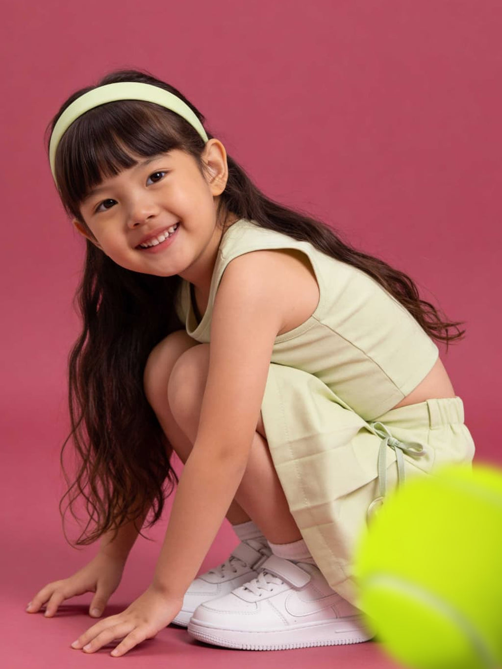 Young girl in a light green dress and white sneakers posing with a tennis ball on a pink background