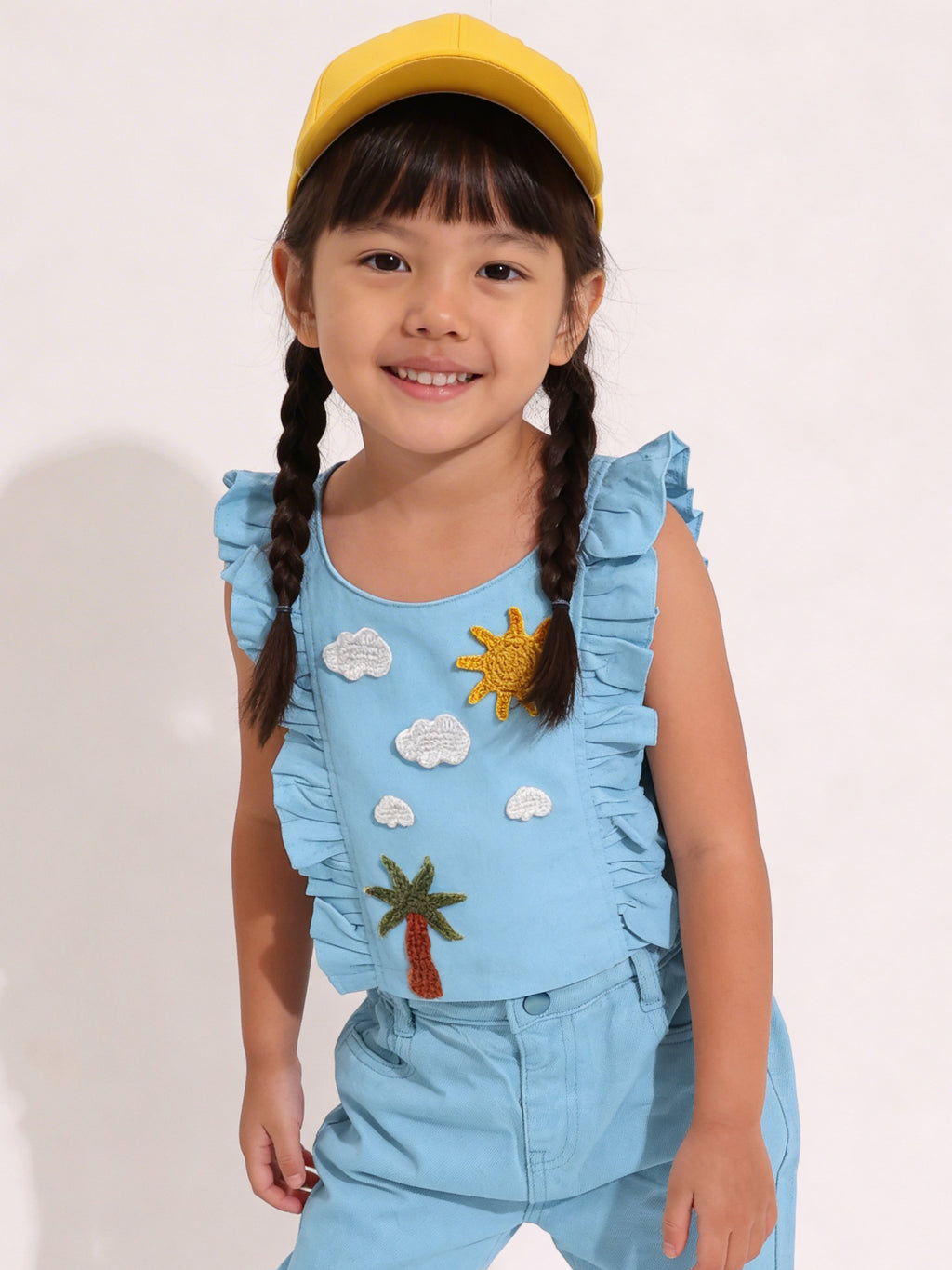 Child wearing a blue Little Carrot outfit with a sun and cloud handmade crochet patches, standing against a plain background