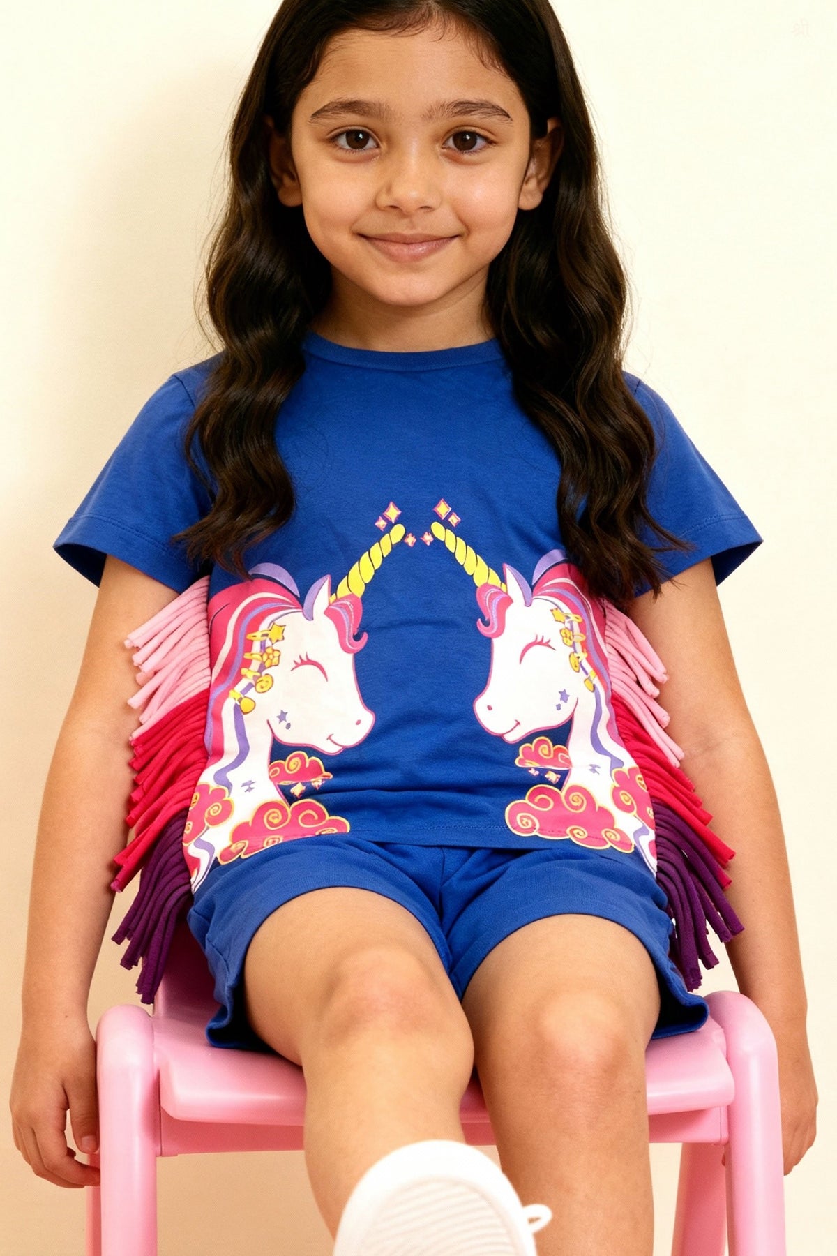 Young girl wearing Little Carrot pure cotton co-ord set with unicorn design and shorts, sitting on a pink chair.