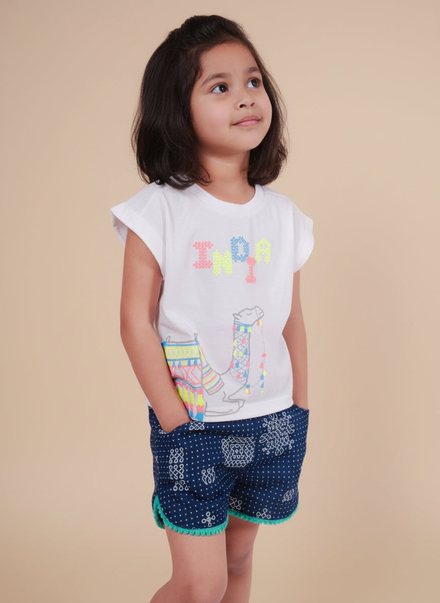 Young girl wearing a white t-shirt with colorful text and blue patterned shorts on a plain background
