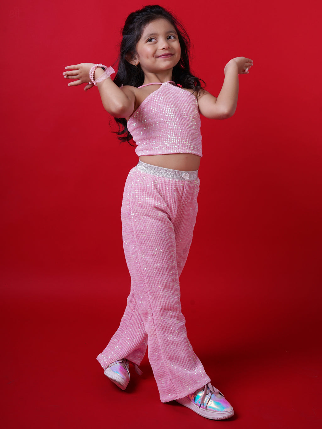 Young girl wearing a Little Carrot pink sequin halterneck top and matching flared pants, smiling with arms raised in a playful pose, standing against a red background.