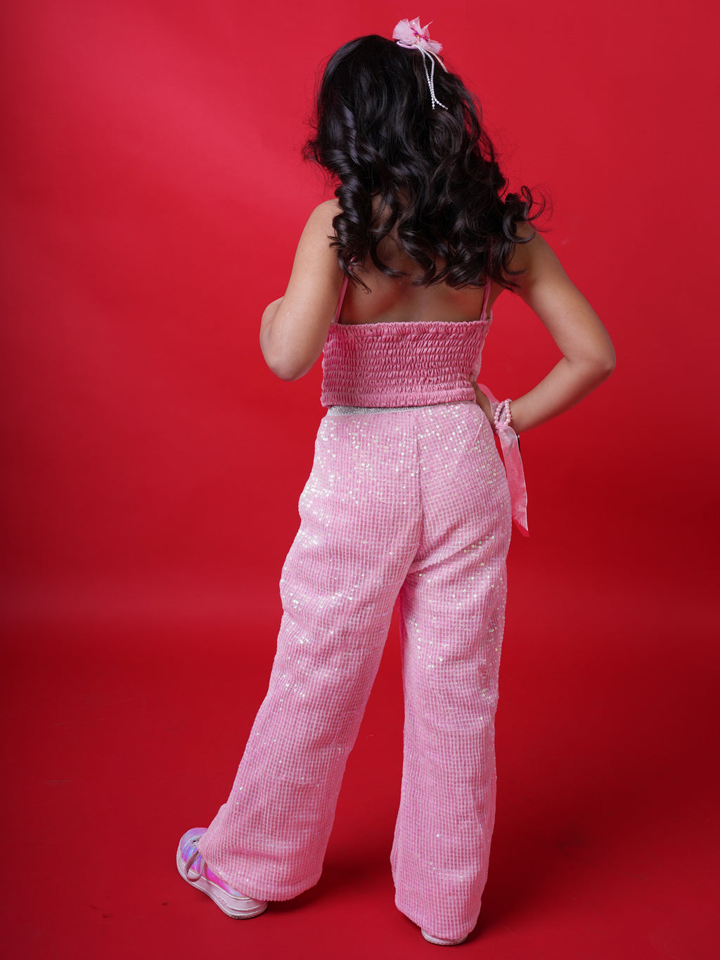 Young girl wearing a Little Carrot pink sequin halterneck top and matching flared pants, smiling with arms raised in a playful pose, standing against a red background.