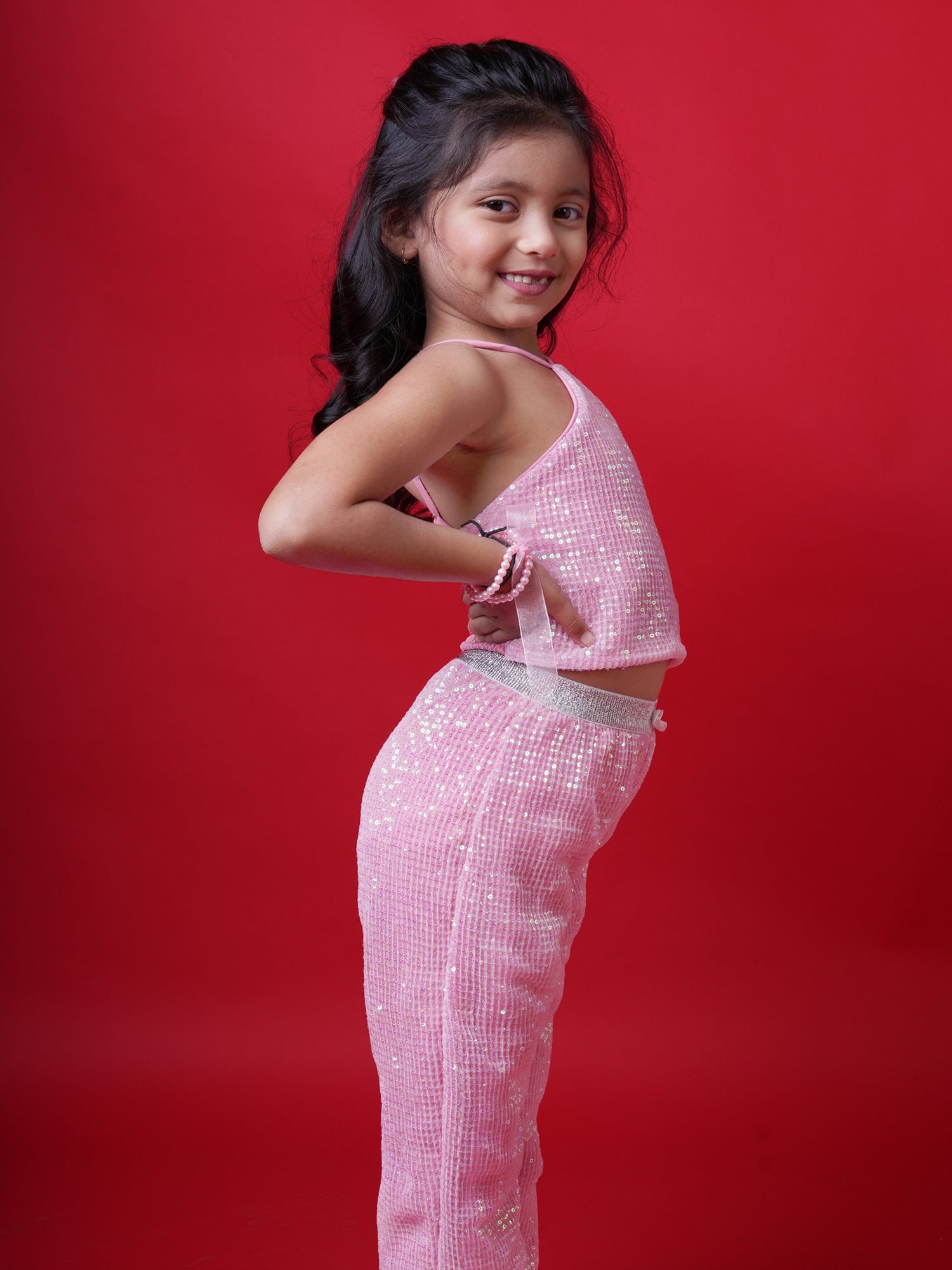Young girl wearing a Little Carrot pink sequin halterneck top and matching flared pants, smiling with arms raised in a playful pose, standing against a red background.