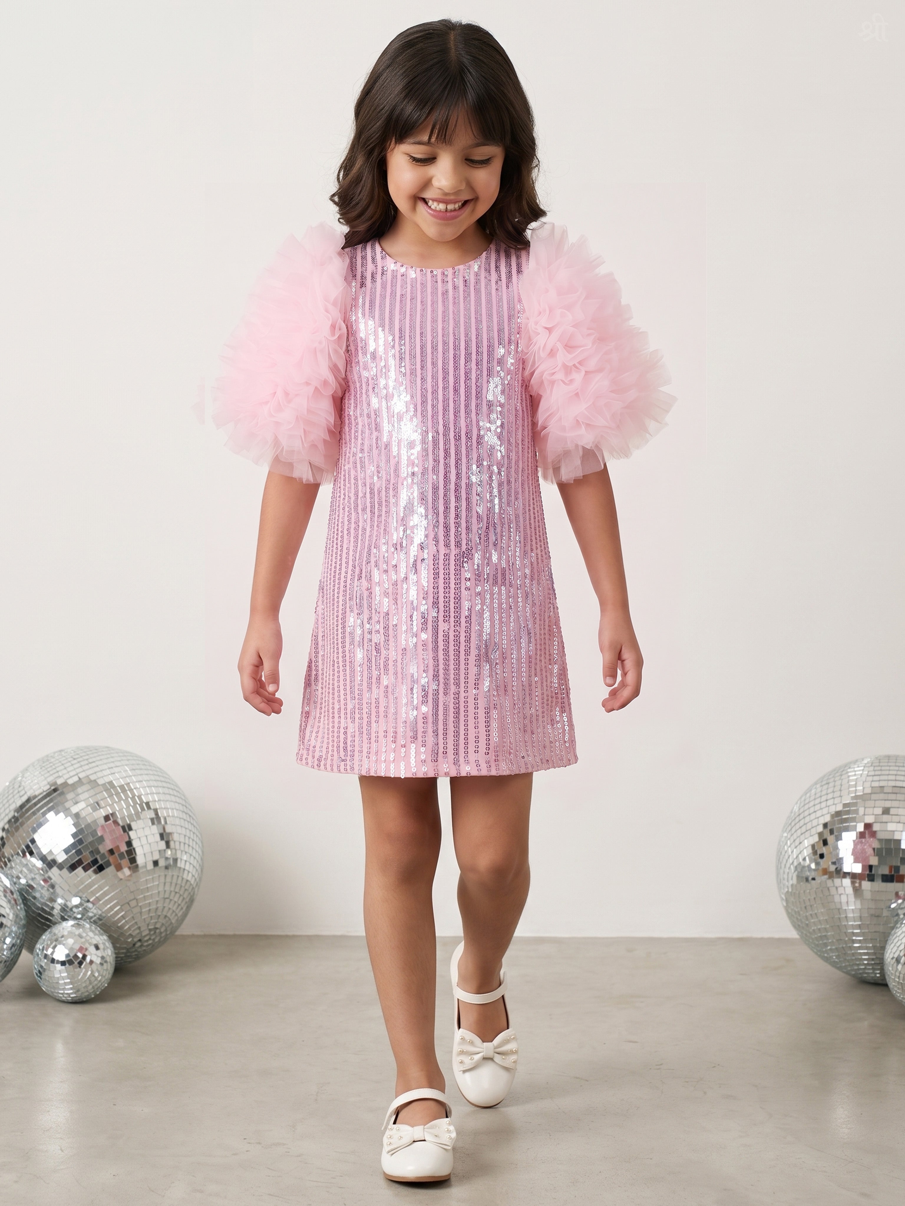 A young girl wearing a Little Carrot pink sequined dress with fluffy tulle puff sleeves, walking in a studio setting with silver disco ball props on the floor.