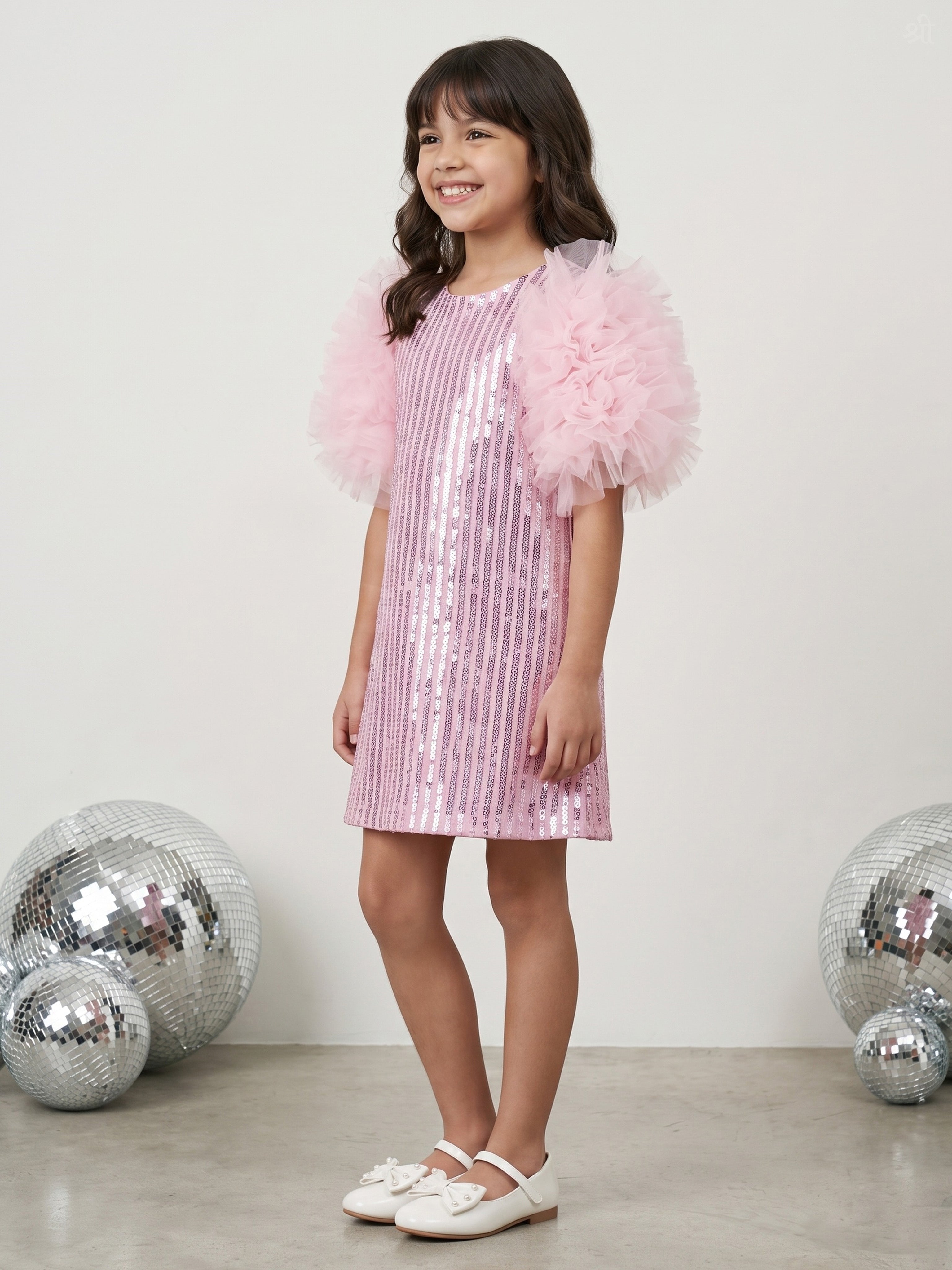 A young girl stands in a studio wearing a Little Carrot light pink sequined dress with fluffy tulle puff sleeves. She is posed in front of a light gray wall with decorative silver disco balls on the floor.