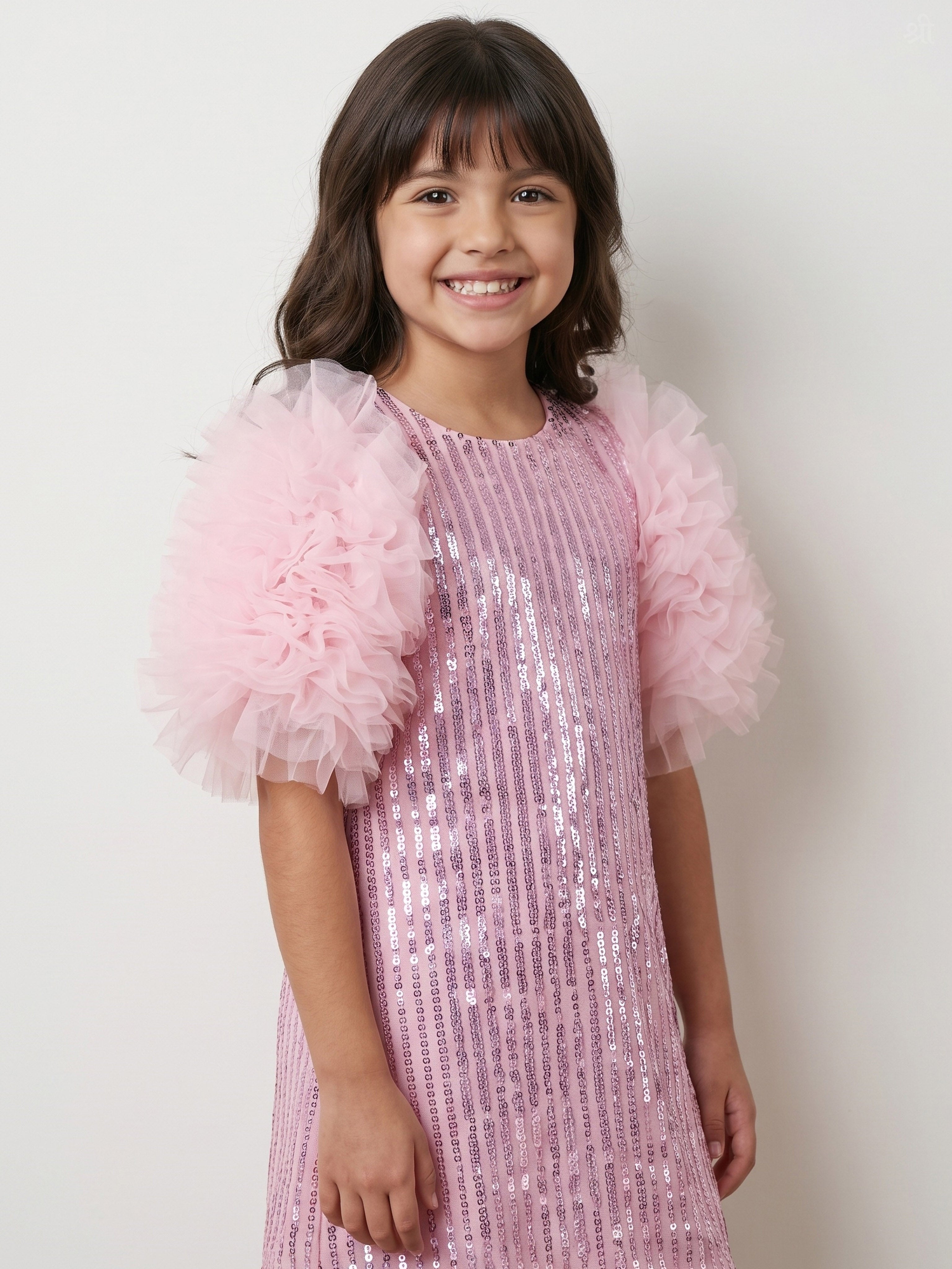 A young girl with dark brown hair smiling, wearing a Little Carrot light pink sequined dress with fluffy tulle puff sleeves, set against a plain off-white background.