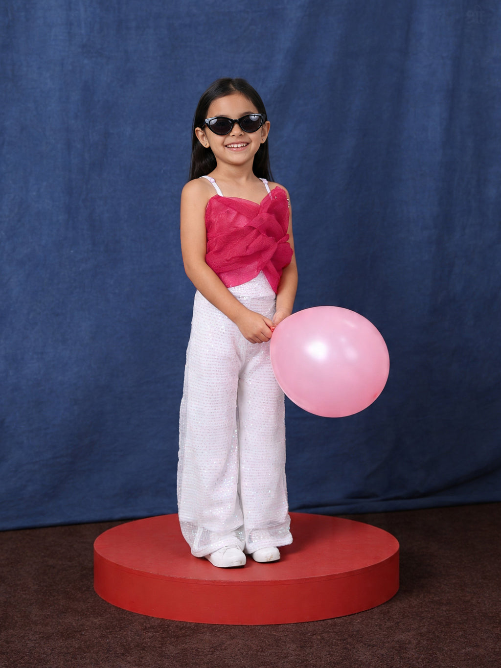 Young girl in a little carrot white sequins jumpsuit with a balloon, standing on a red platform against a blue background.