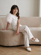 A Young girl happily sitting on couch in a Little Carrot White Sequins Jumpsuit