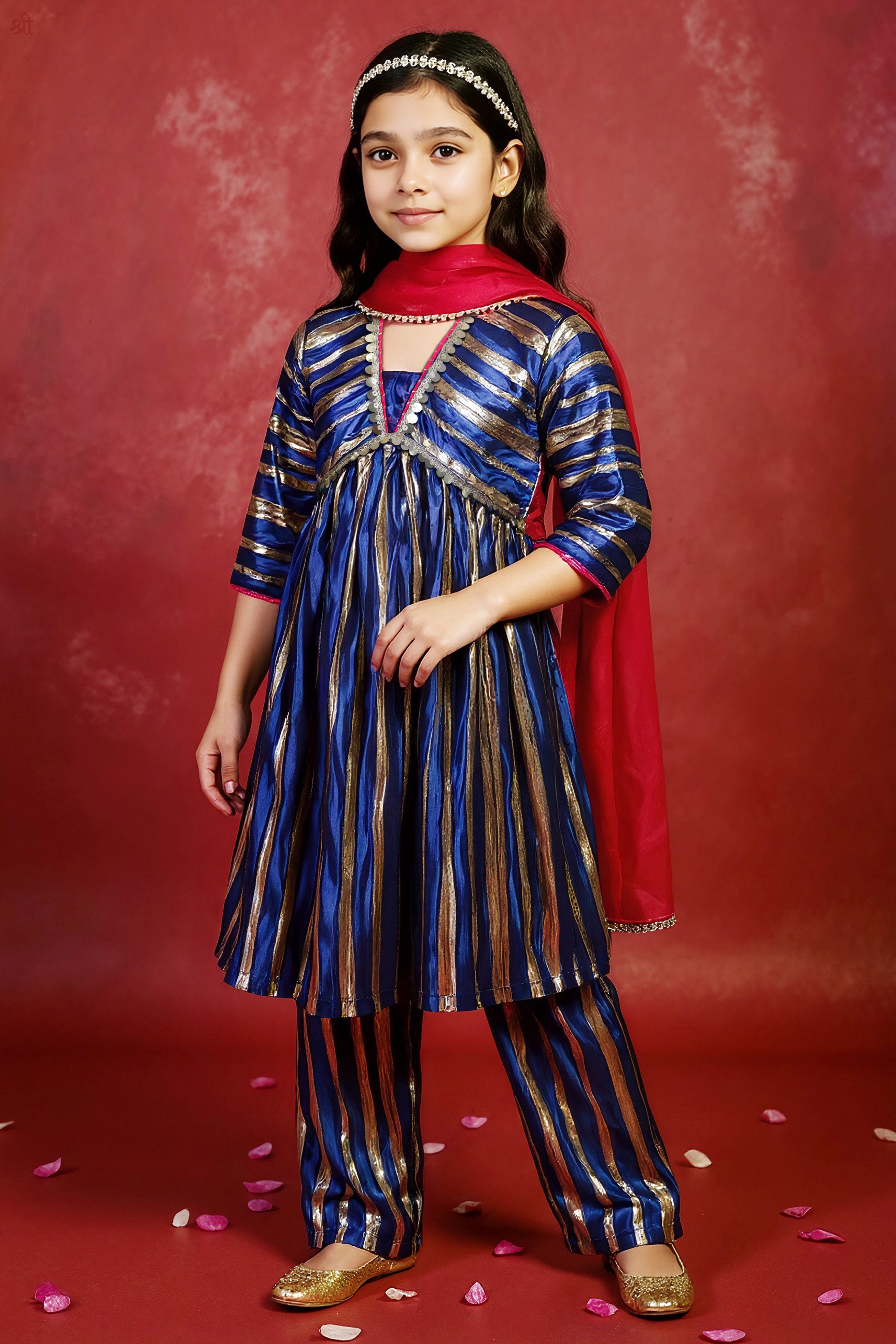 Young girl in a traditional little carrot blue and gold outfit with a fuchsia dupatta against a red background.