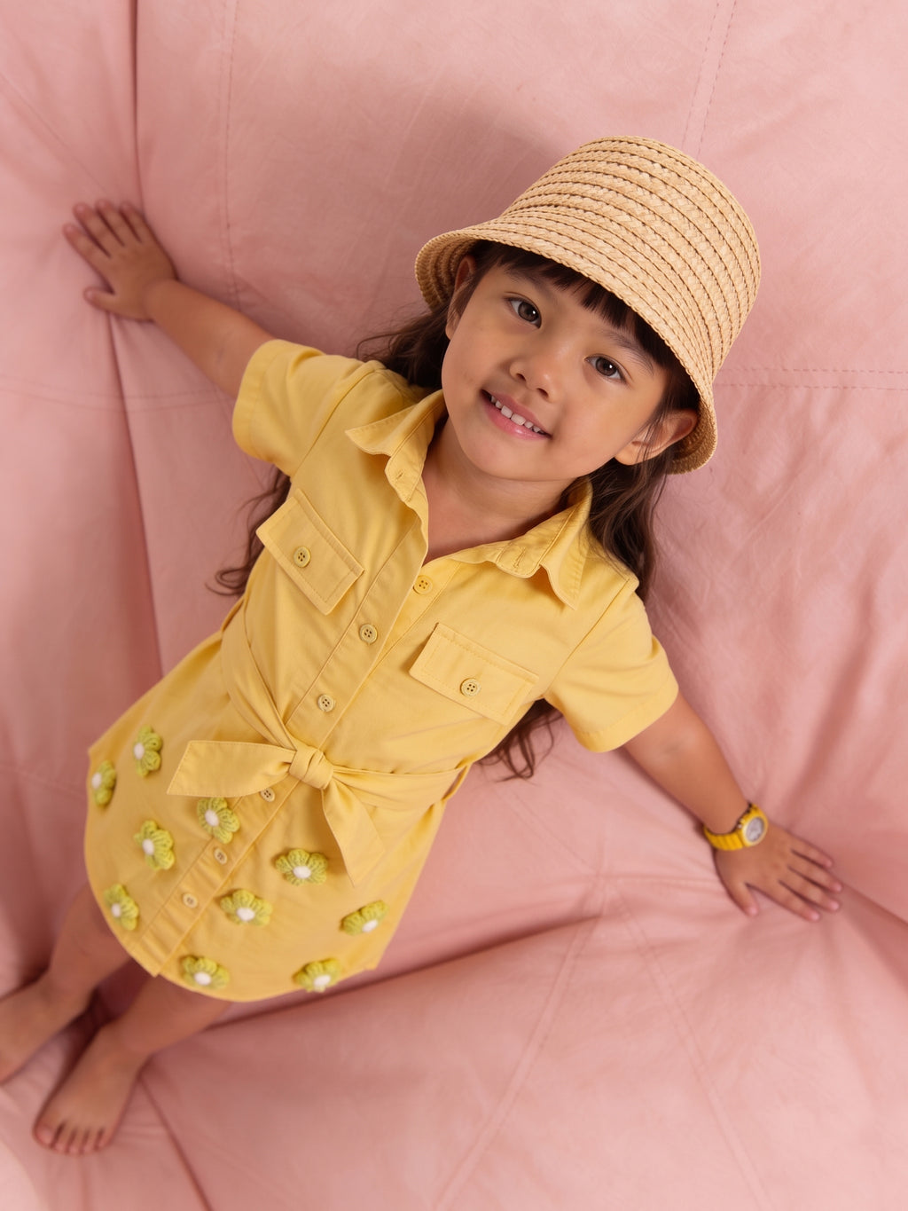 Child wearing a yellow little carrot dress with a bow and a straw hat on a pink background