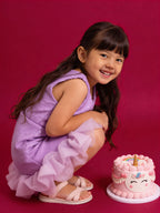 Young girl in a Little Carrot purple Sequins dress with ruffles sitting next to a unicorn-themed cake on a red background
