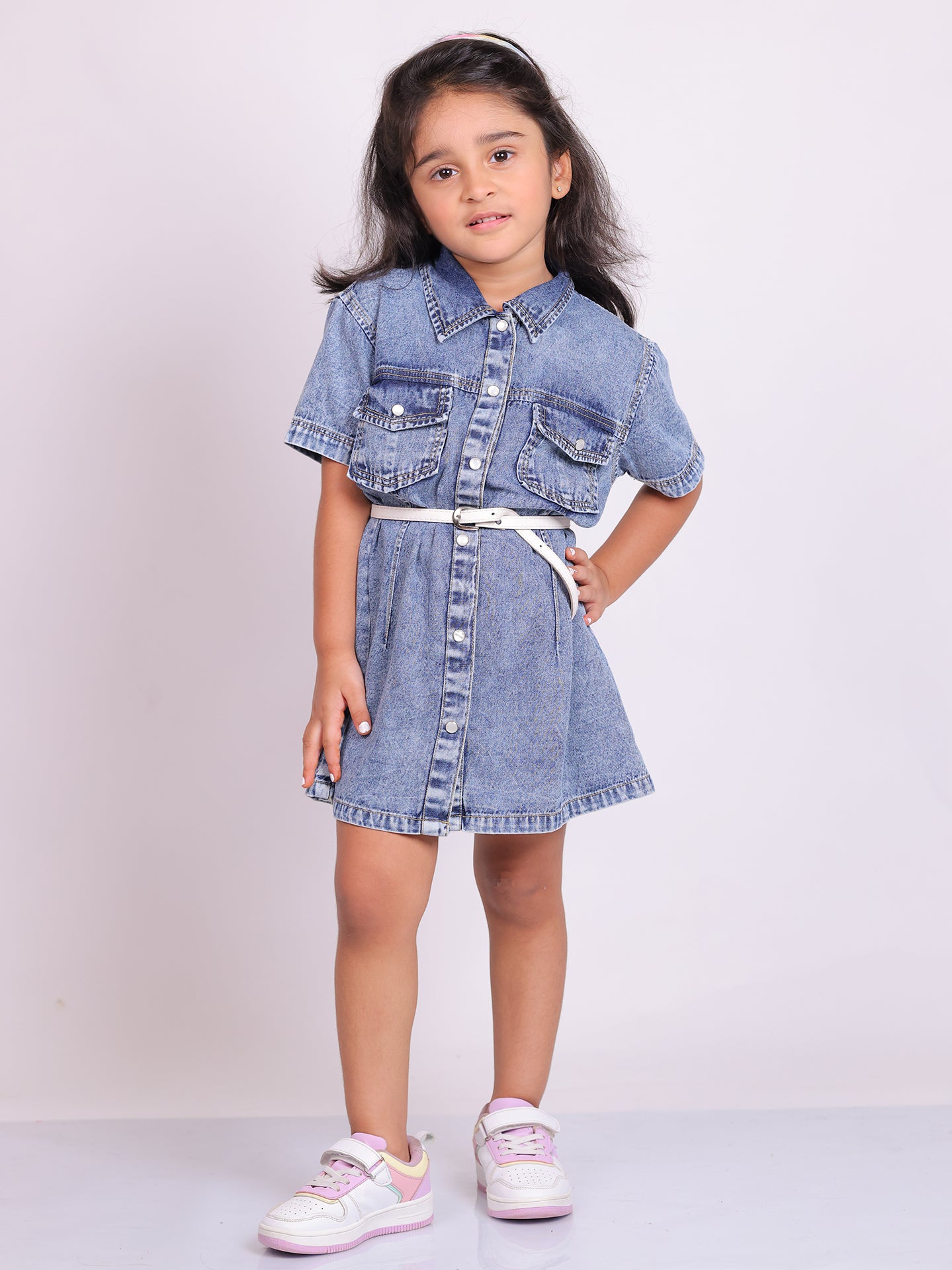 A young girl wearing a stylish denim shirt dress with short sleeves, snap buttons, and two chest pockets. The knee-length dress is cinched at the waist with a white belt, creating a structured yet comfortable fit. She pairs the look with trendy white and pink sneakers, posing confidently against a plain white background. This premium kidswear piece is ideal for casual outings and semi-formal occasions. Available for B2B wholesale and white-label manufacturing by Sundaram Fashions.