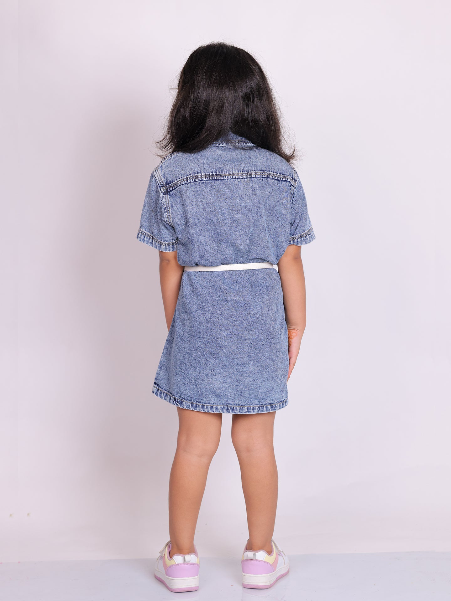 A young girl wearing a stylish denim shirt dress with short sleeves, snap buttons, and two chest pockets. The knee-length dress is cinched at the waist with a white belt, creating a structured yet comfortable fit. She pairs the look with trendy white and pink sneakers, posing confidently against a plain white background. This premium kidswear piece is ideal for casual outings and semi-formal occasions. Available for B2B wholesale and white-label manufacturing by Sundaram Fashions.