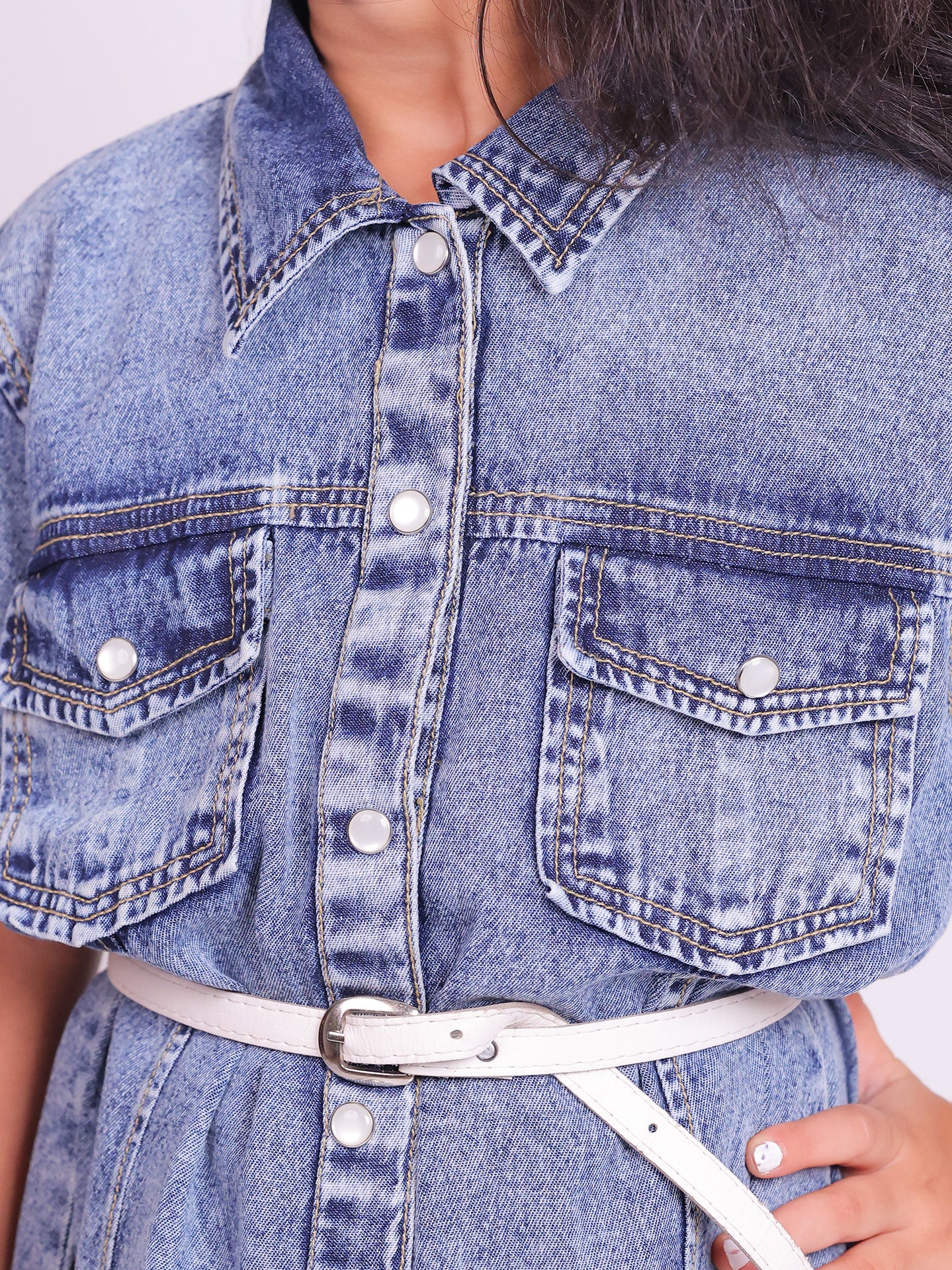 A young girl wearing a stylish denim shirt dress with short sleeves, snap buttons, and two chest pockets. The knee-length dress is cinched at the waist with a white belt, creating a structured yet comfortable fit. She pairs the look with trendy white and pink sneakers, posing confidently against a plain white background. This premium kidswear piece is ideal for casual outings and semi-formal occasions. Available for B2B wholesale and white-label manufacturing by Sundaram Fashions.
