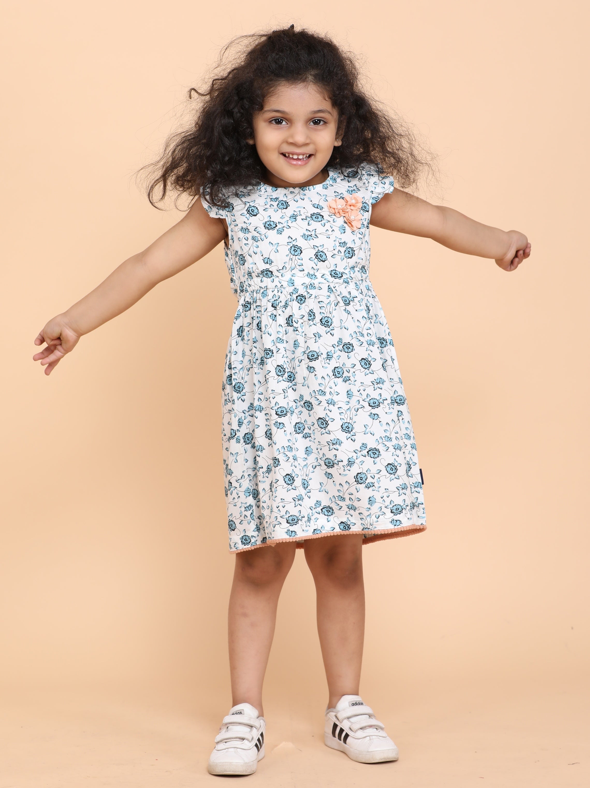 Smiling girl wearing a white floral summer dress with frill sleeves and handmade flower detail at the chest, designed by Little Carrot.